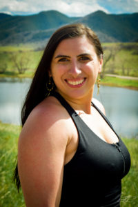 Woman with black hair wearing a black tank top while standing in front of a mountain lake.