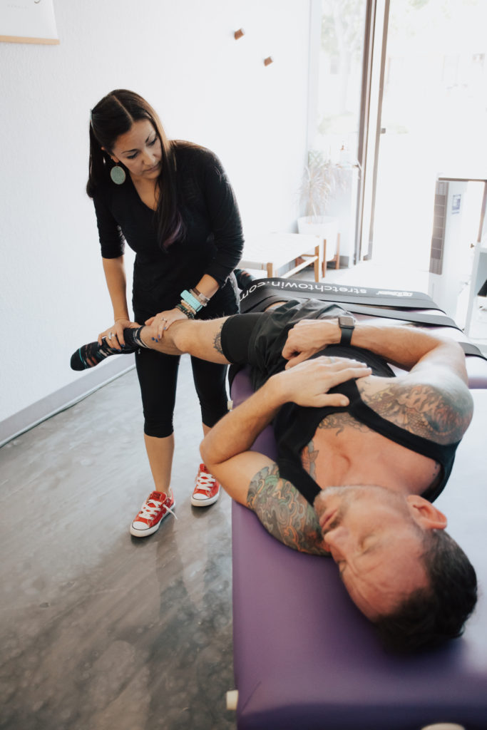 Athletic man with tattoos laying on a purple stretch table.  He is being stretched by a stretch therapist wearing all black
