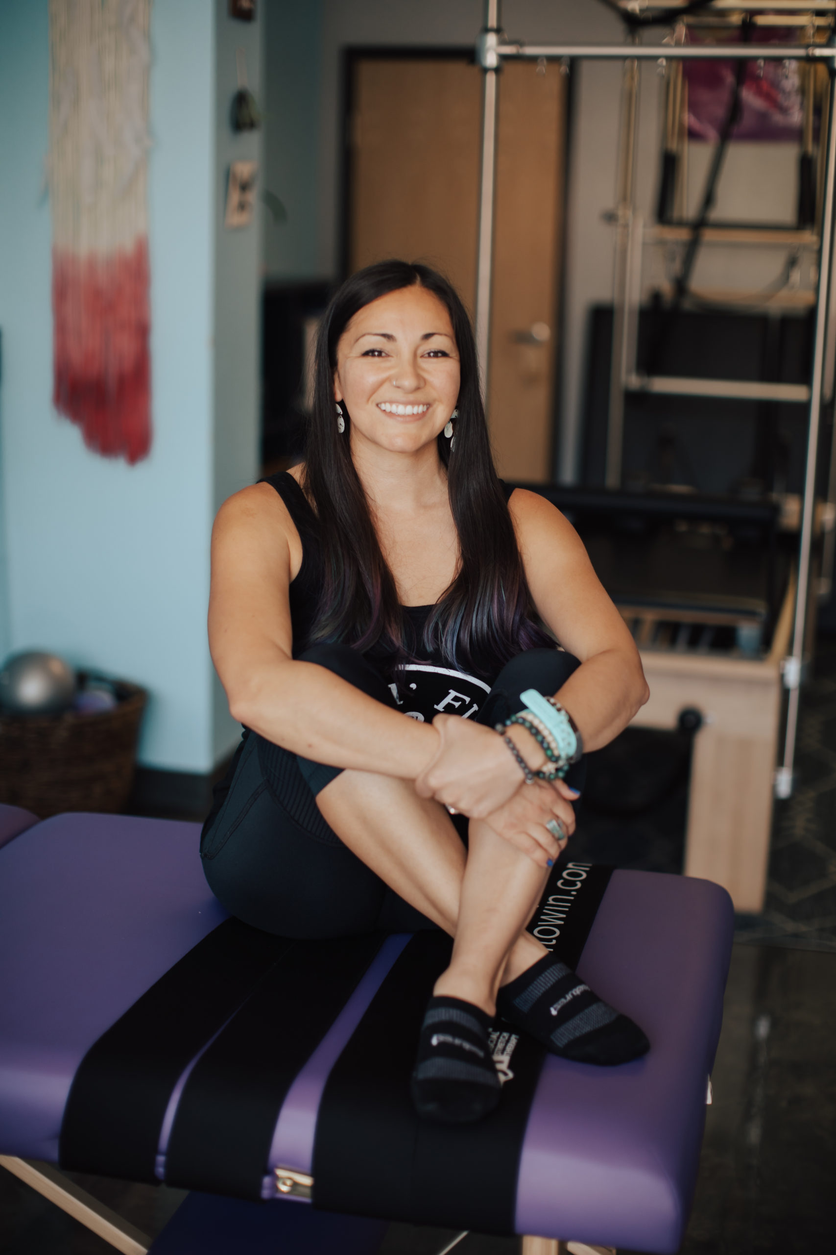 Pilates instructor wearing all black, sitting crossed legged on a purple stretch table.