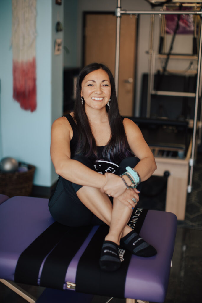 Pilates instructor wearing all black, sitting cross legged on a purple stretch table.
